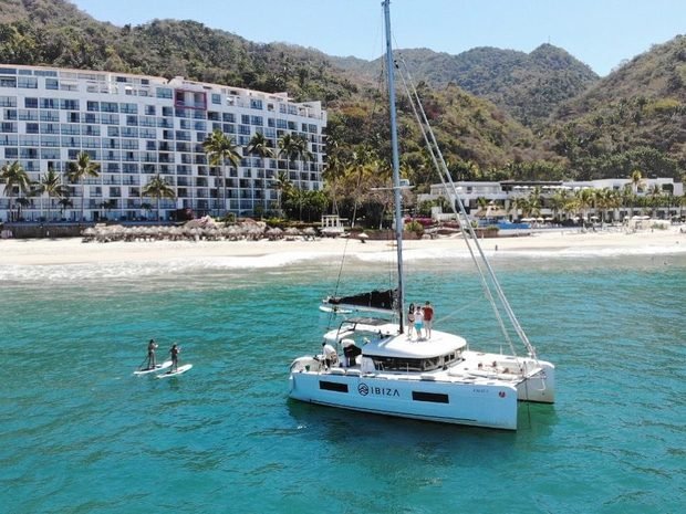 A catamaran at anchor off a beach, with paddleboarders alongside